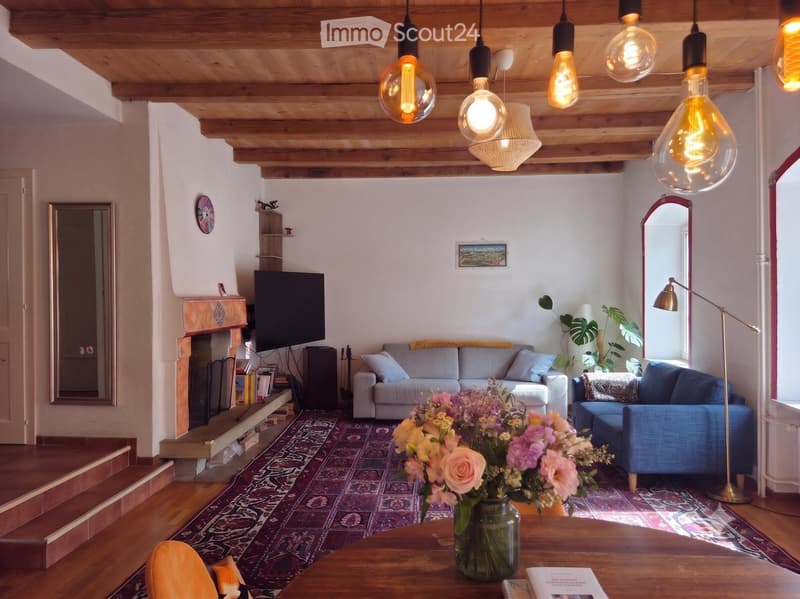 Living room with exposed wooden beams, a fireplace, a sofa, a chair, a coffee table with a flower vase, and pendant lights.