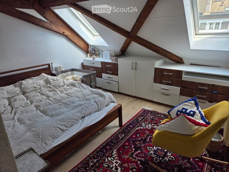 Cozy attic bedroom with wooden beams, skylight, bed, yellow armchair, patterned rug, desk, and built-in cabinets.