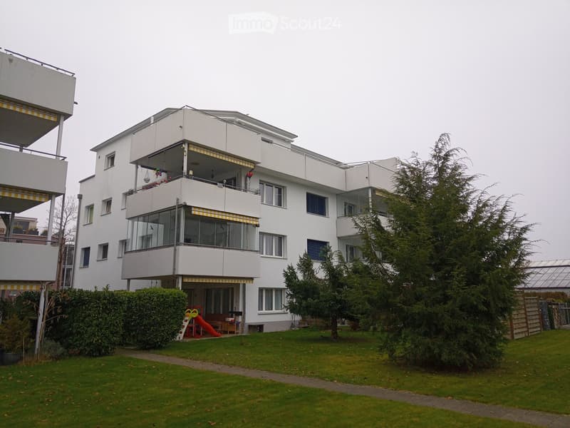 Modern three-story residential building with white facade, multiple balconies, large windows, and surrounding greenery including trees and shrubs.