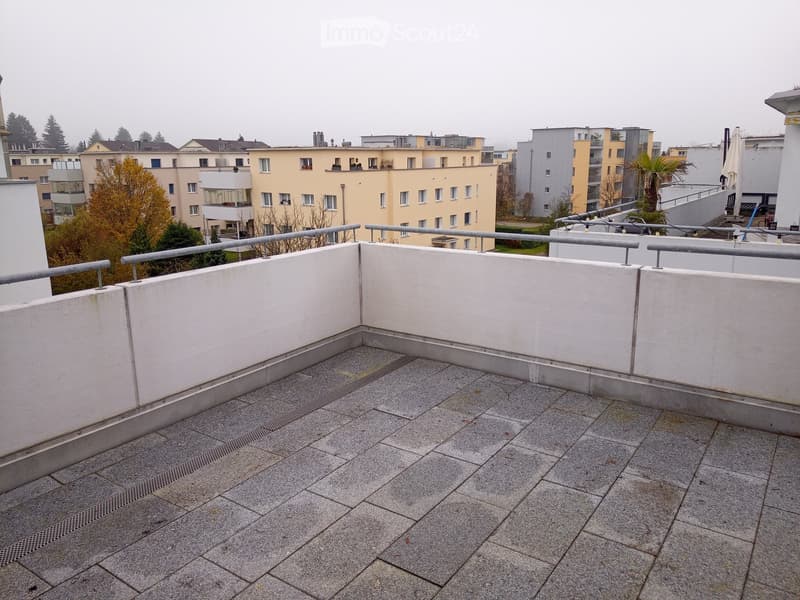 Empty rooftop terrace with grey tiled flooring, white low walls, and metal railings, overlooking a residential area with buildings and trees.