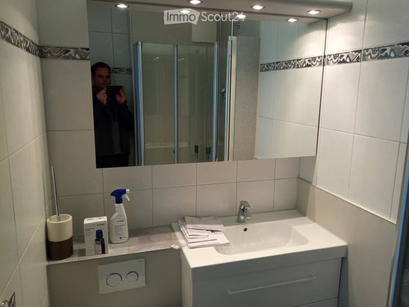 Modern bathroom with white tiles, a large mirror with built-in lighting, a sink with a modern faucet, and various toiletries on the counter.