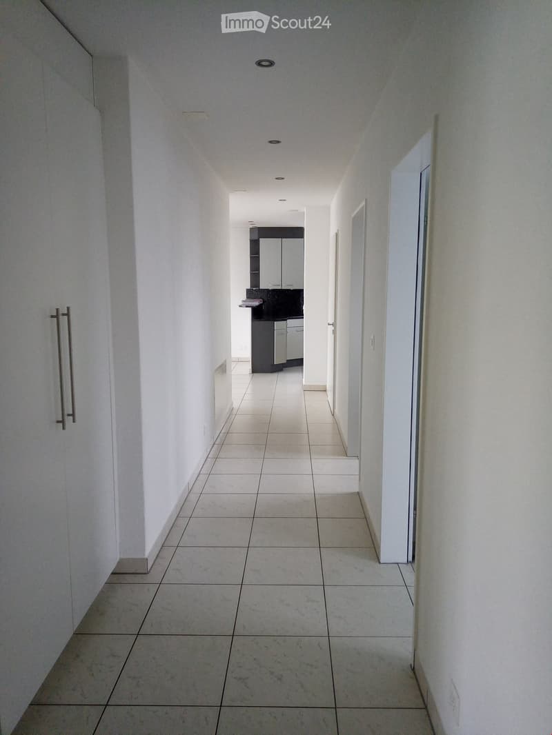 A clean, modern hallway with white tiled flooring and walls, featuring recessed ceiling lights and a small kitchen area visible at the end.