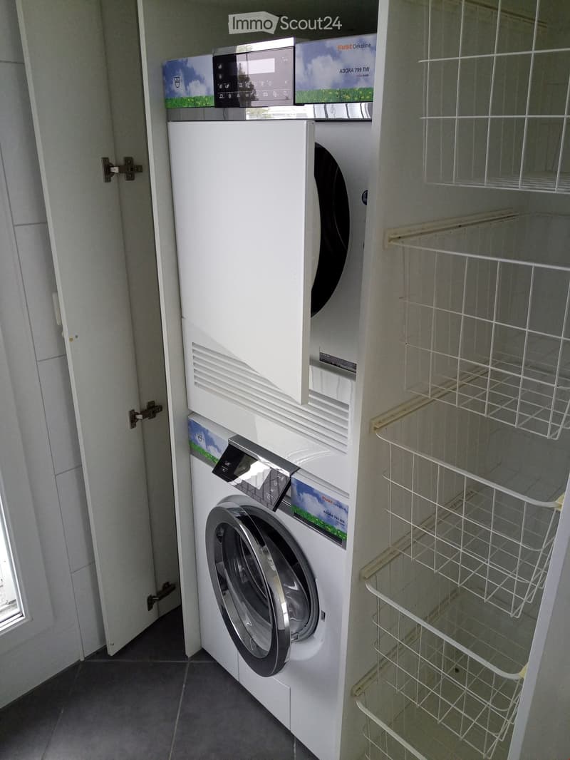 Modern bathroom with a white washing machine and dishwasher stacked vertically, accompanied by white wire shelves on the right side.