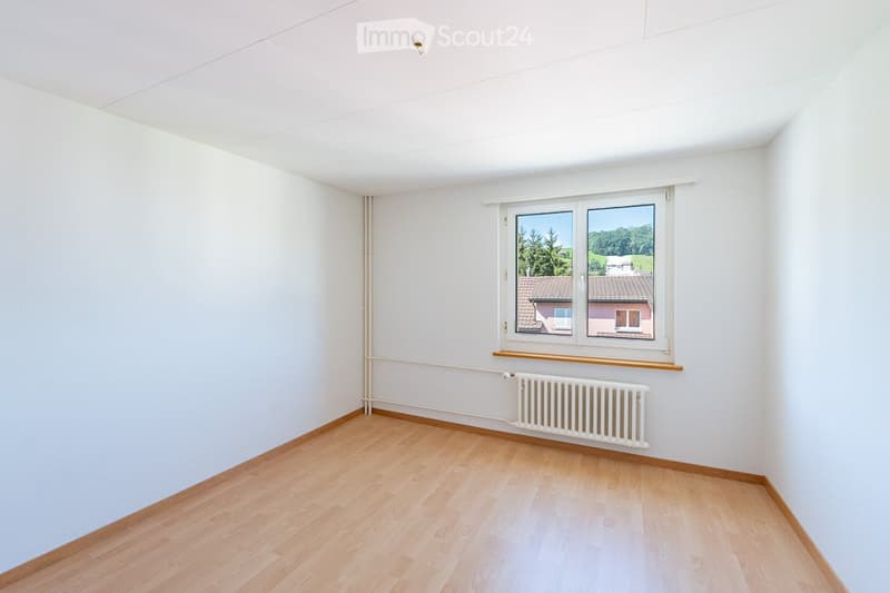 Empty room with light wooden flooring, white walls, and a single window offering a view of the outdoors.