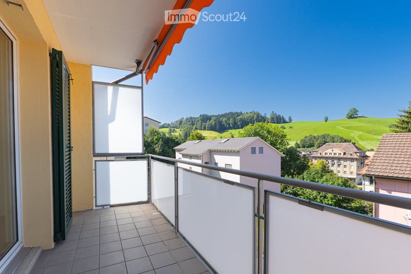 Balcony with tiled floor, metal railing, and glass panels, overlooking a scenic countryside with green hills and houses.