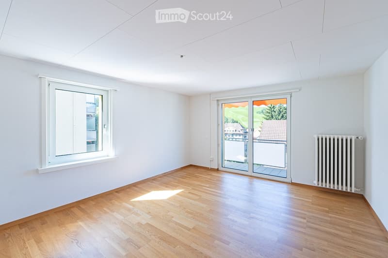Empty room with wooden flooring, white walls, large window, and sliding glass door leading to a balcony.