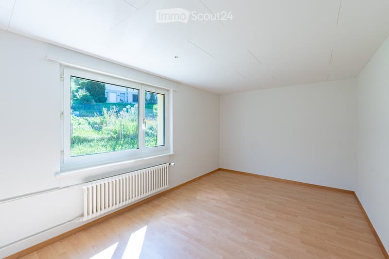 Empty room with white walls, wooden flooring, and a large window showing greenery outside. No furniture or equipment is present.