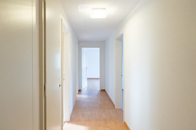 A clean, well-lit hallway with wooden flooring, white walls, and several doors leading to different rooms.