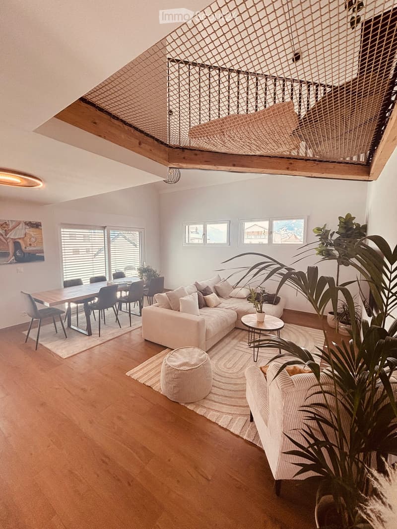 Open-plan living and dining area with light-colored wood flooring, a sectional sofa, coffee table, ottoman, dining table with chairs, and large windows with blinds.