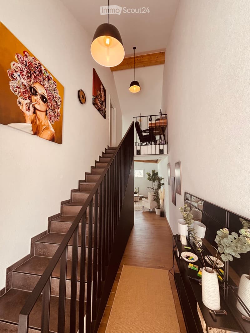 Modern hallway with wooden flooring, staircase, pendant lights, and artwork on the walls. Features a small table with plants and decorative items.