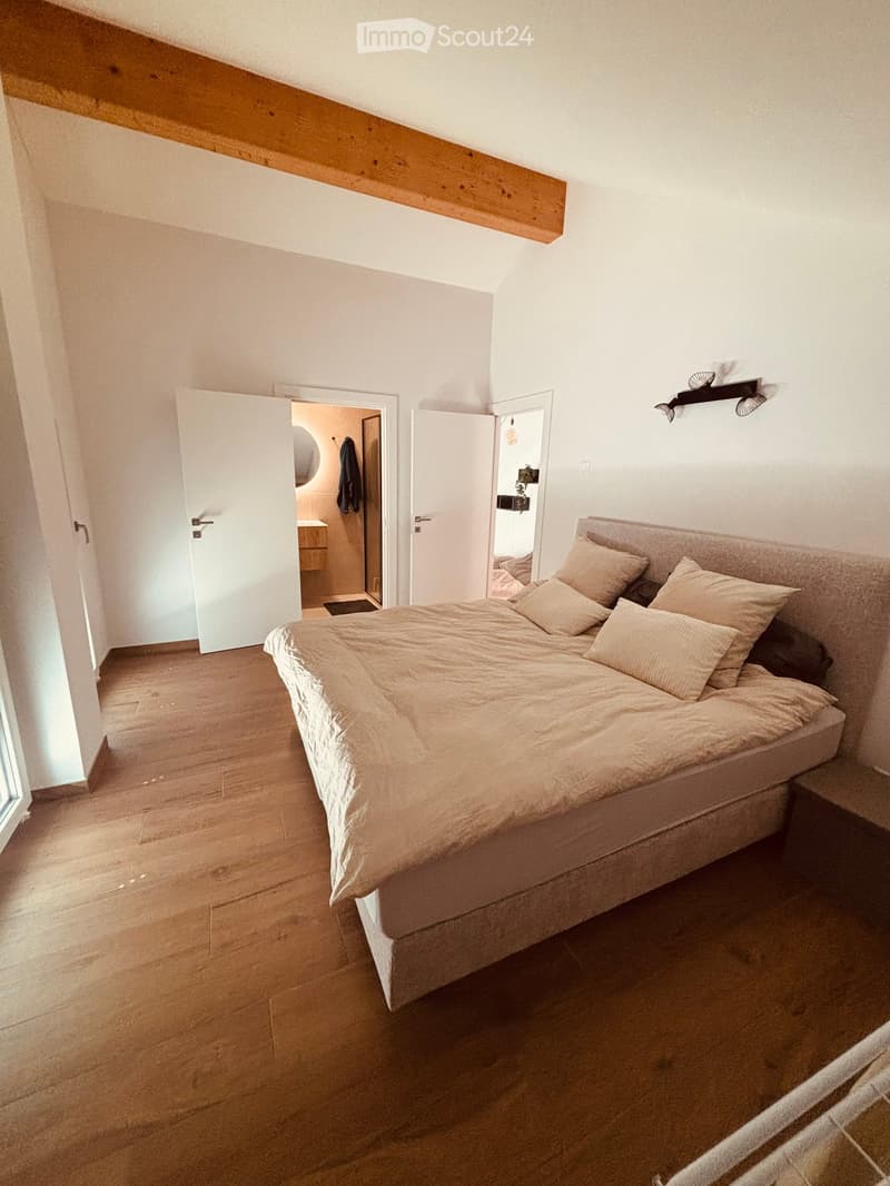 Cozy bedroom with a large bed, light-colored bedding, and wooden flooring. The room features an open doorway leading to a bathroom and another room.