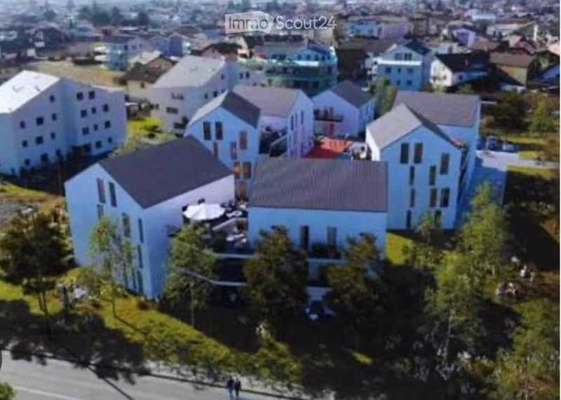 Aerial view of a residential area with multiple modern, light blue houses with grey roofs, surrounded by greenery and trees.