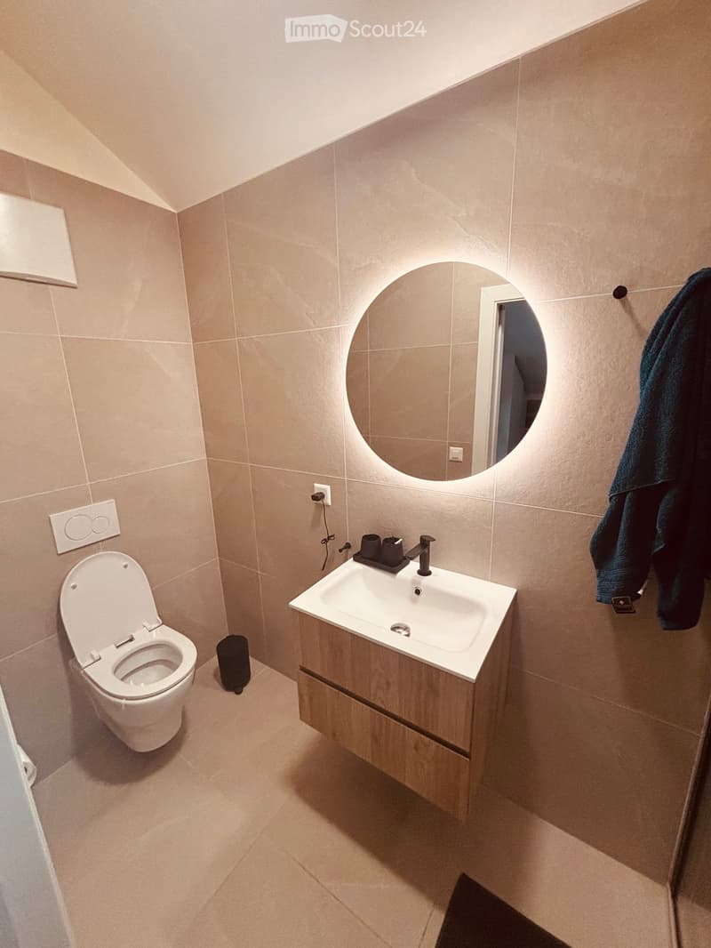 Modern bathroom with beige tiles, a white toilet, a sink with a modern faucet, a round mirror with built-in lighting, and a towel hanging on the wall.