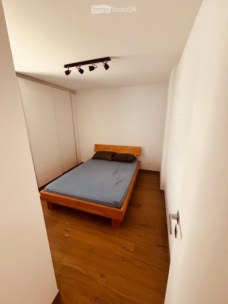 A small bedroom with a wooden bed, blue mattress, and two pillows, featuring wooden flooring and white walls.