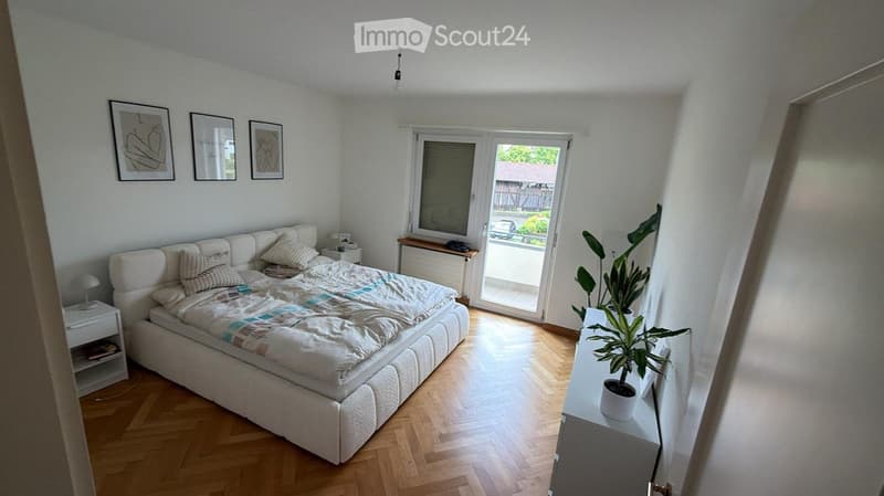 Bright bedroom with wooden flooring, large bed with white headboard and bedding, nightstand, plant, and artwork on the wall. Large window with blinds and a view of the outdoor area.
