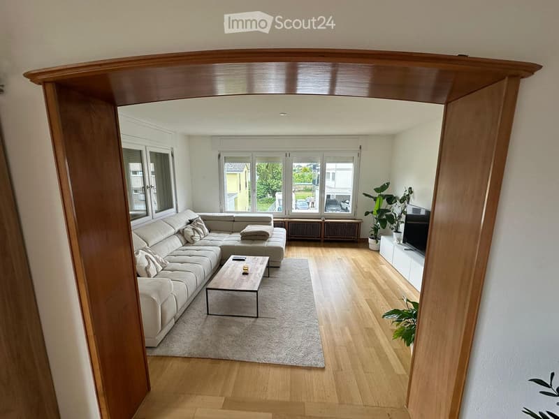 Modern living room with light-colored sectional sofa, coffee table, TV, and plants. Large windows provide natural light.