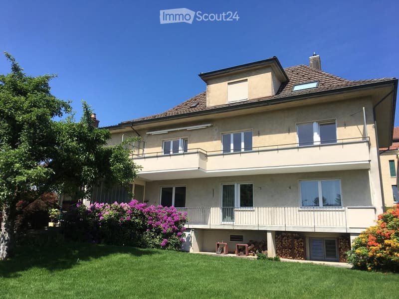 Two-story house with beige exterior, multiple windows, balconies, and a well-maintained lawn with colorful flowering shrubs.