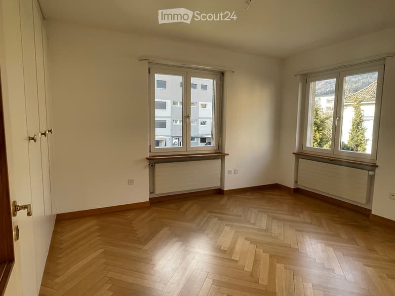 Empty room with wooden herringbone flooring, white walls, and large windows with radiators beneath them. The room has a door on the left side.