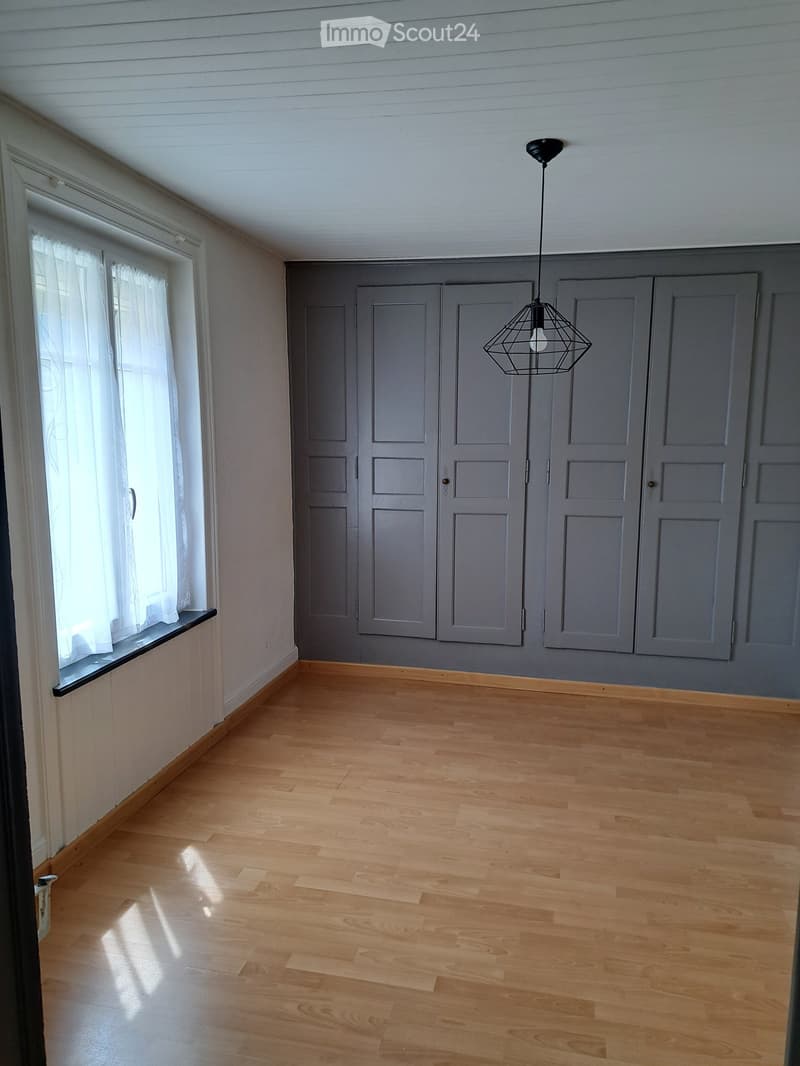 Empty room with wooden flooring, grey walls, large window with white curtains, and grey cabinetry.