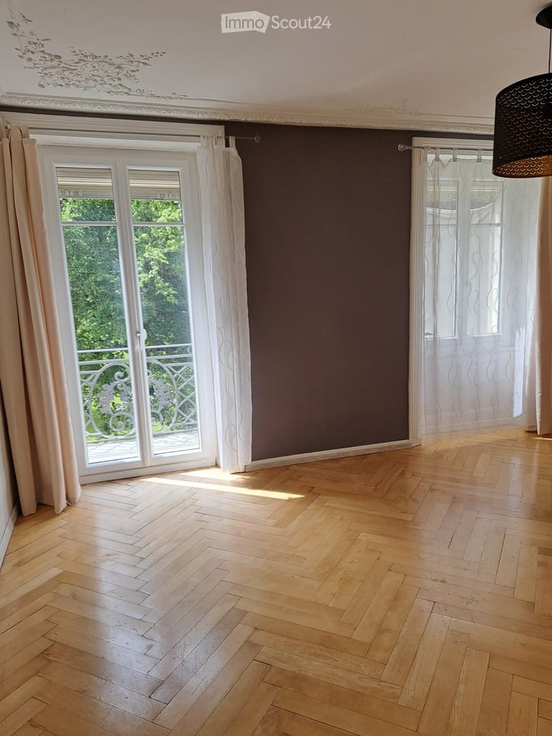 Empty room with wooden herringbone flooring, dark brown accent wall, white curtains, and large windows with a view of greenery.