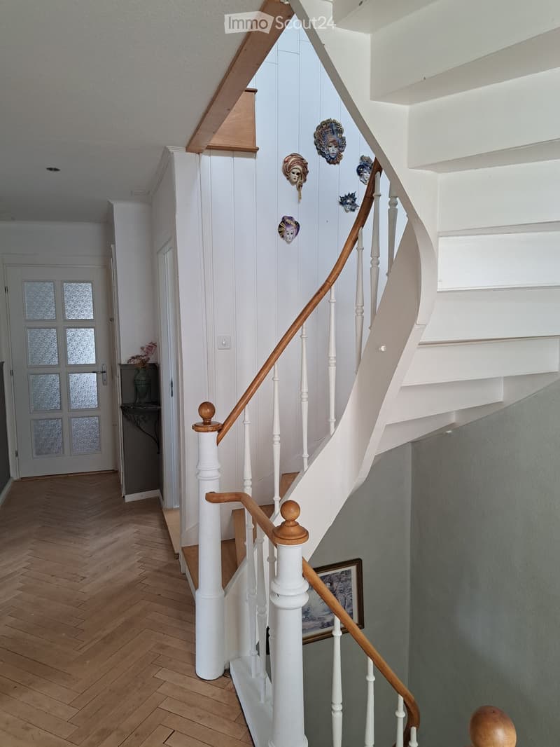 Spiral staircase with white balusters and wooden handrail, decorative masks on wall, wooden herringbone floor, framed picture on wall, small table with vase and flowers near entrance door.