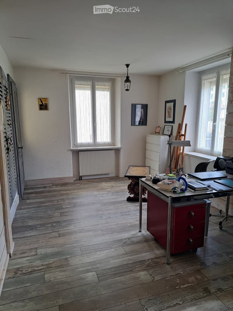 Office space with wooden flooring, large windows, a desk with office supplies, a chair, a cabinet, a small table with a framed picture, and a radiator under the window.