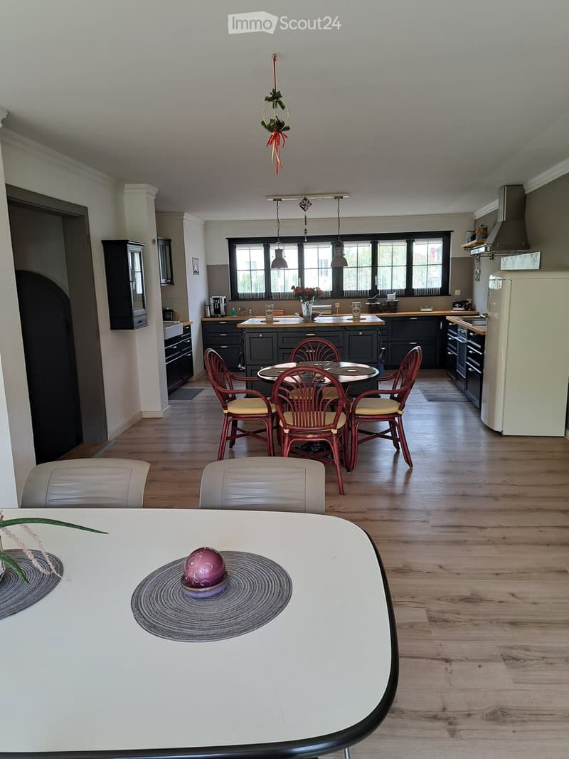 Bright kitchen with dark cabinets, stainless steel appliances, and a dining area with a round table and chairs. The space features wooden flooring, pendant lights, and a large window with blinds.
