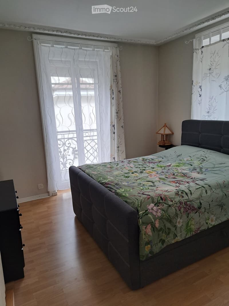Bedroom with wooden flooring, large bed with floral duvet, nightstand with table lamp, and sheer curtains on windows.