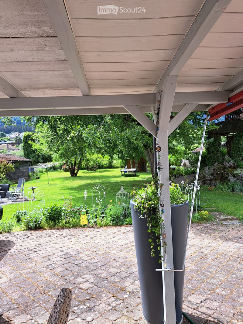A covered outdoor patio area with a stone-paved floor, large potted plants, and a view of a well-maintained lawn and garden area with trees and decorative elements.