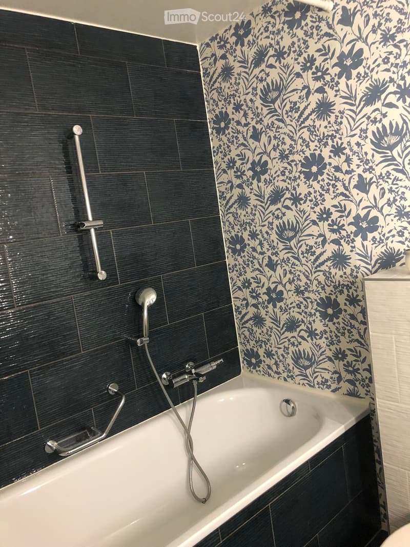 Bathtub with modern dark tiles and floral patterned wall, equipped with a handheld showerhead and a handrail.