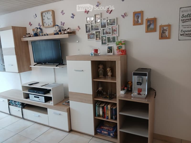 A modern living room with a television on a wooden shelf, a coffee machine on a side table, books on a bookshelf, and decorative items including picture frames and figurines.