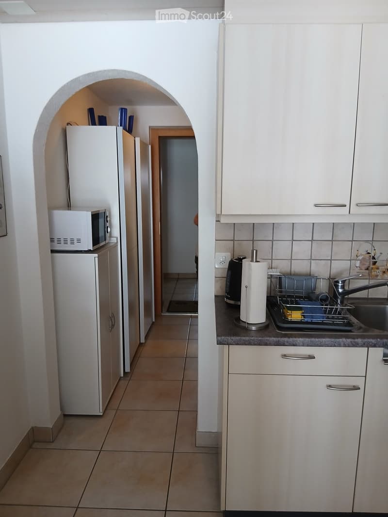 The image shows a compact kitchen area with white cabinets, a refrigerator, microwave, sink, and various small appliances. The floor is tiled, and the space features an arched entryway leading to another room.