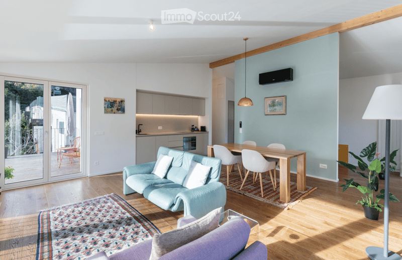 Modern living room with a light blue sofa, wooden dining table, white chairs, kitchen area, large windows, and a balcony view.
