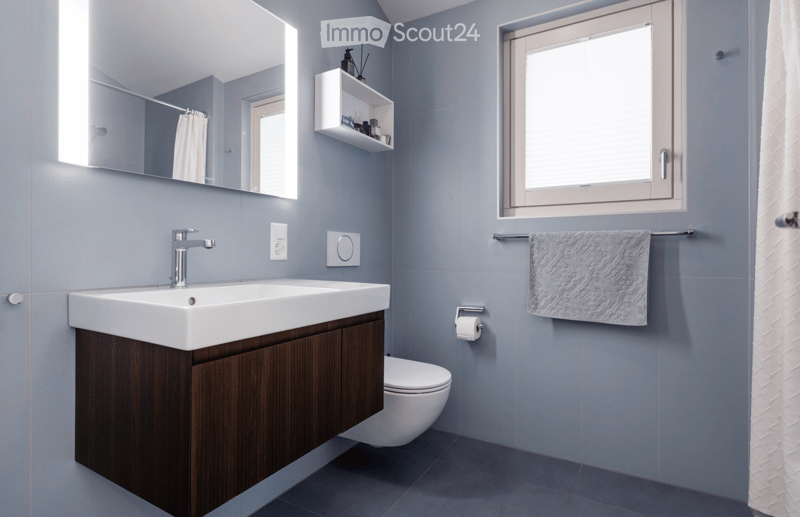 Modern bathroom with a white sink, dark wooden cabinet, chrome faucet, large mirror, and a window. Includes a towel on a rack, a shelf with toiletries, and a toilet.