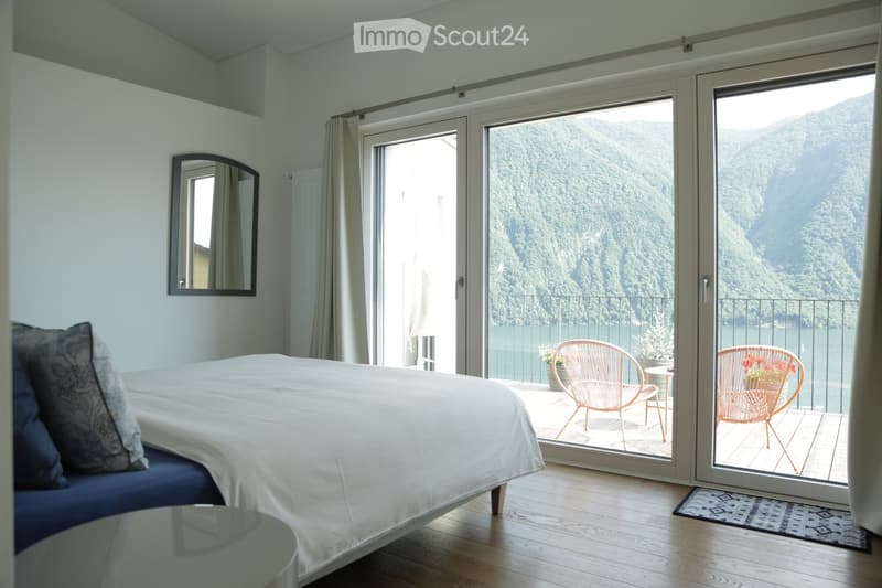 Modern bedroom with a large bed, a mirror on the wall, and a balcony overlooking a scenic lake view. The balcony features two chairs and a small table.