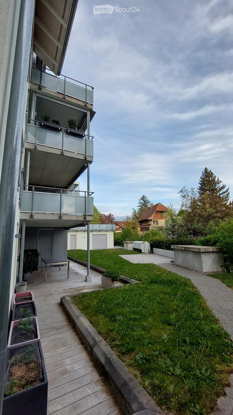 Modern outdoor area with wooden decking, raised planters, small table, and grassy lawn. Adjacent to a multi-story building with balconies and glass railings.