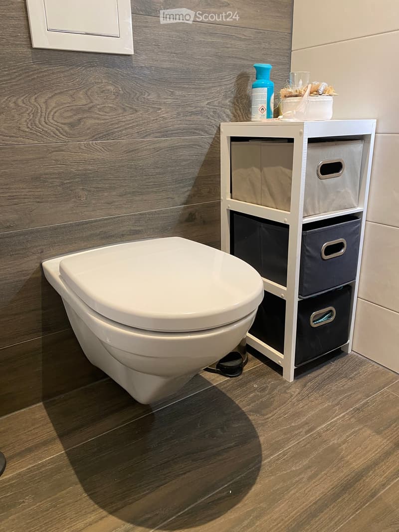 Modern bathroom with a white toilet, wooden floor, and a white storage unit with multiple compartments. A bottle and a cup are placed on top of the storage unit.