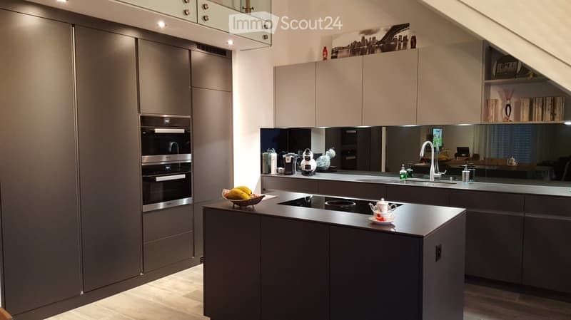 Modern kitchen with dark grey cabinets, stainless steel appliances, island with black countertop, and various kitchen items including a teapot, bowl of fruit, and coffee maker.