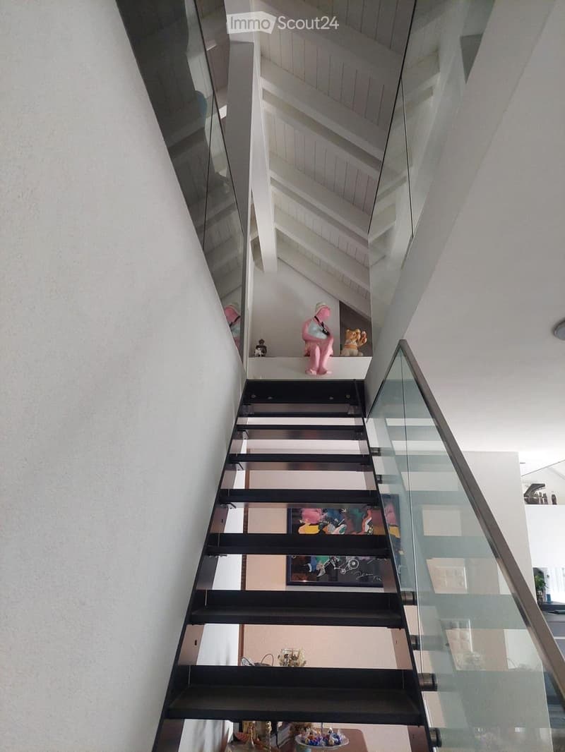 Modern staircase with black steps and glass railing, leading to an open-plan living area with visible artwork and decorative items on shelves.