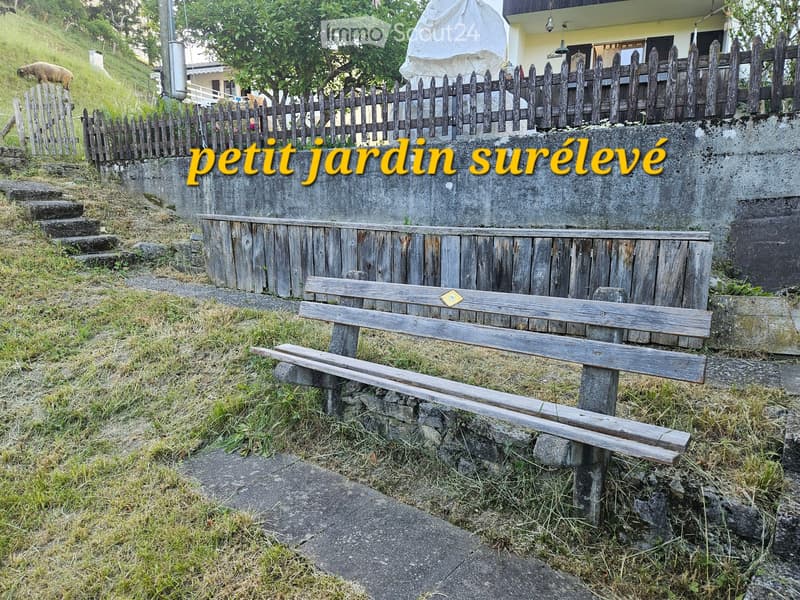 A small garden area with a wooden bench placed on a stone platform, surrounded by a wooden fence and grassy hillside.