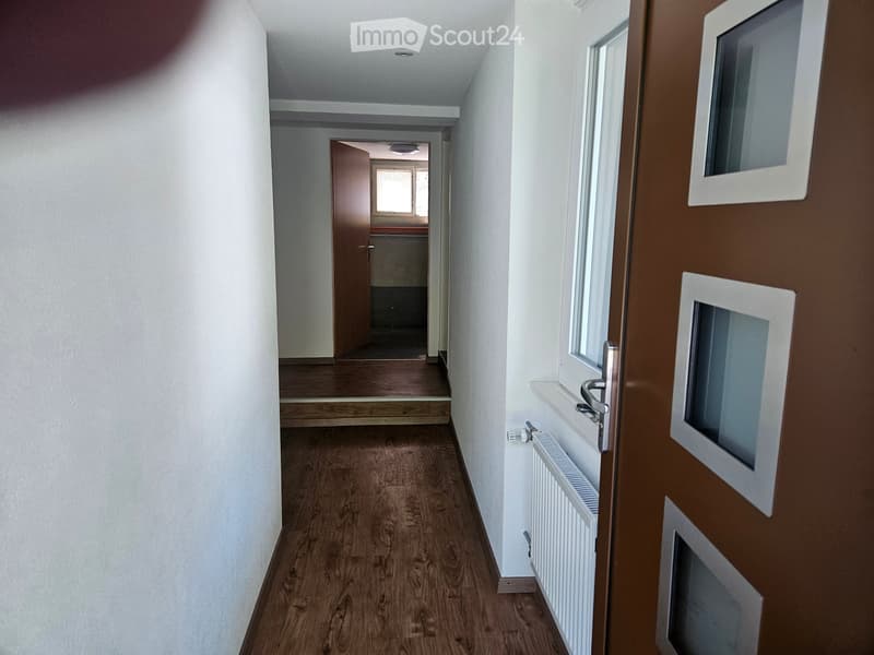 Empty hallway with wooden flooring, white walls, and a radiator on the right side. A door at the end leads to another room with visible furniture.