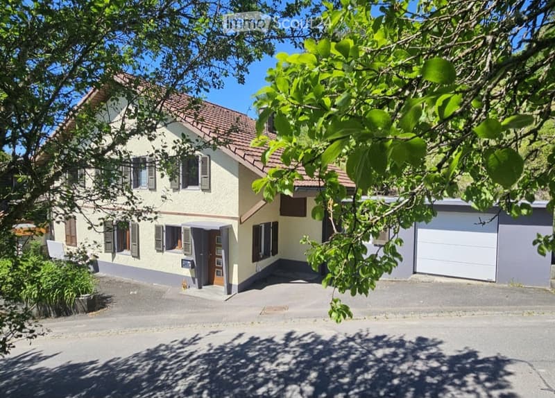 Two-story house with beige walls, red-tiled roof, multiple windows with shutters, and a garage attached to the right side.