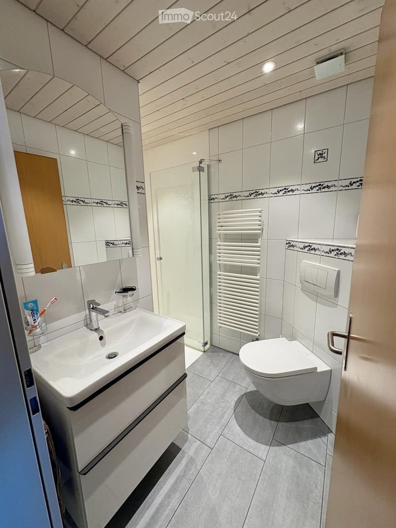 Modern bathroom with white tiles, a sink with a modern faucet, a toilet, a glass-enclosed shower area, and a white radiator. The walls feature decorative tiles with a floral pattern near the base.