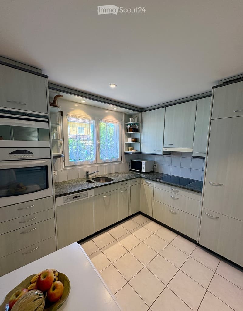 Modern kitchen with light-colored cabinets, granite countertops, and stainless steel appliances including an oven, dishwasher, and microwave. A bowl of fruit is placed on the counter.