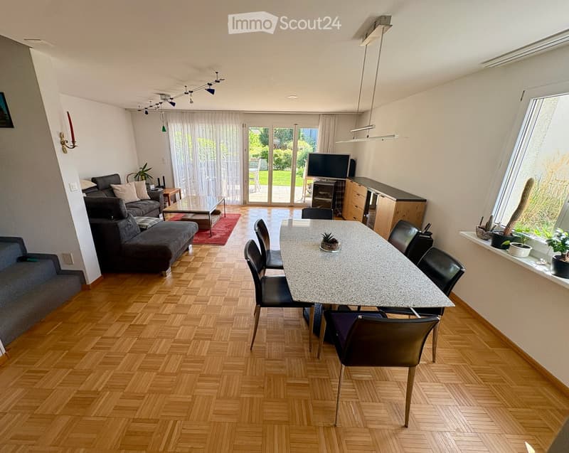 Open-plan living and dining area with wooden flooring, large windows, and sliding glass doors leading to a garden. Features a grey sectional sofa, coffee table, dining table with chairs, TV, and decorative plants.
