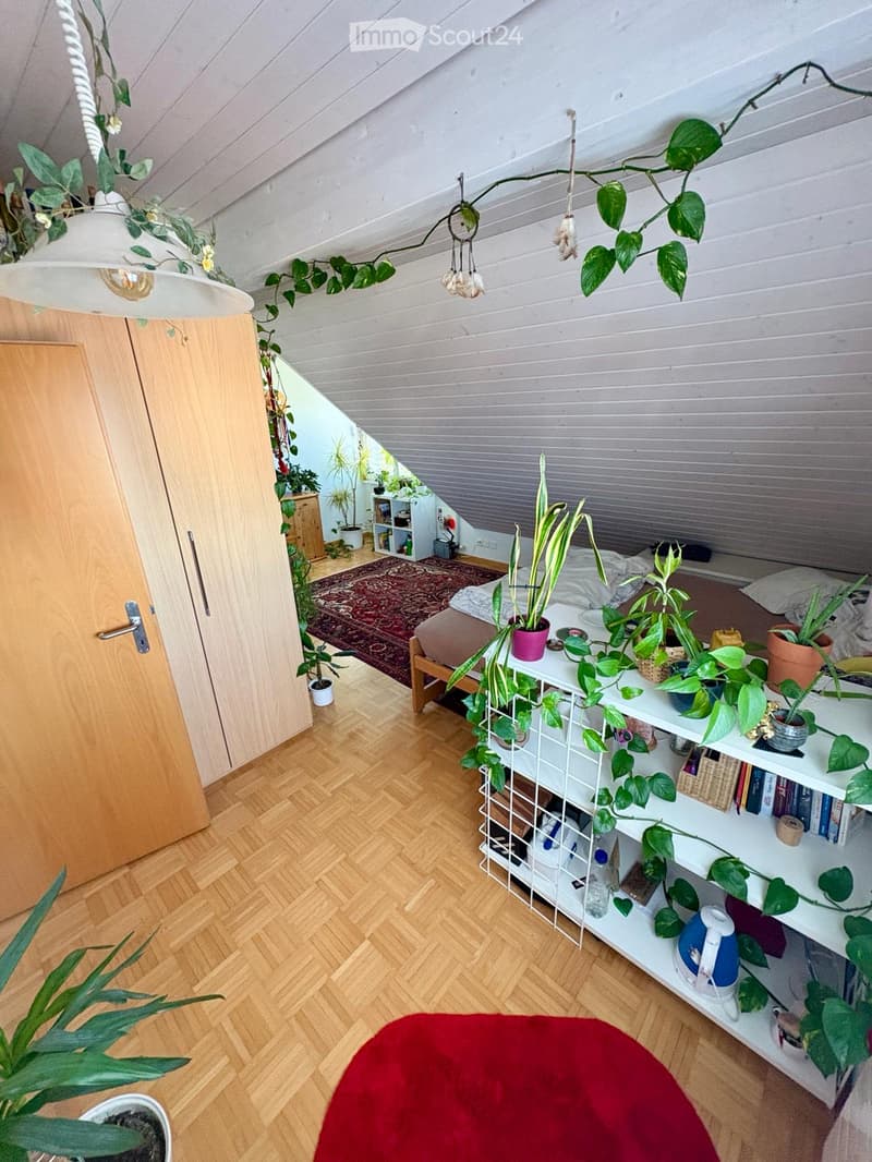 A cozy attic bedroom with wooden floors, slanted ceilings, and multiple potted plants. Features a bed, a shelf with books and plants, and a hanging plant arrangement.