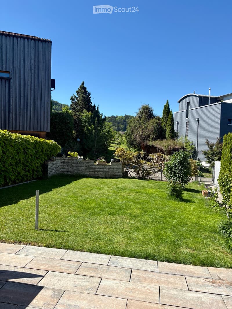 Well-maintained garden with a large lawn, stone pathway, low stone wall, and surrounding trees and shrubs.