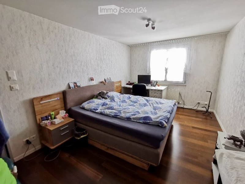 A cozy bedroom with a wooden floor, a large bed with a patterned duvet, a nightstand, a desk with a computer, and a window with white curtains.