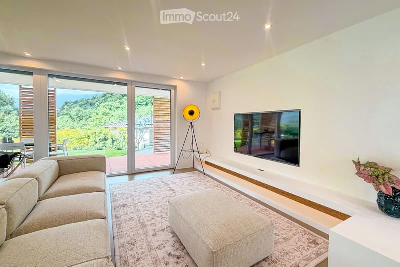 Living Room. Modern living room with large sliding glass doors offering a scenic view, a grey sectional sofa, ottoman, flat-screen TV, floor lamp, and a small table with a plant.