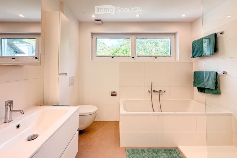 Bathroom. Modern bathroom with a white sink, toilet, bathtub, and large windows offering a scenic view. Green towels are neatly hung on a rack, and the space is well-lit with recessed ceiling lights.